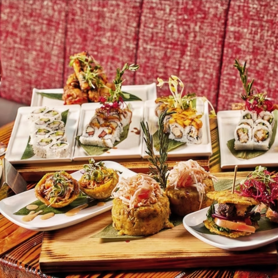 A vibrant spread of Latin-American dishes, including sushi rolls, plantain cups, and mofongo, on a wooden board.